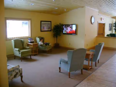 A cozy senior living lounge with green armchairs, a wall TV, plants, and a reception area.