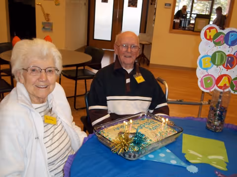 Two elderly individuals sitting at a table with a birthday cake that has lit candles. There is a colorful 'Happy Birthday' sign on the table. The setting appears to be a communal area in a senior living facility.