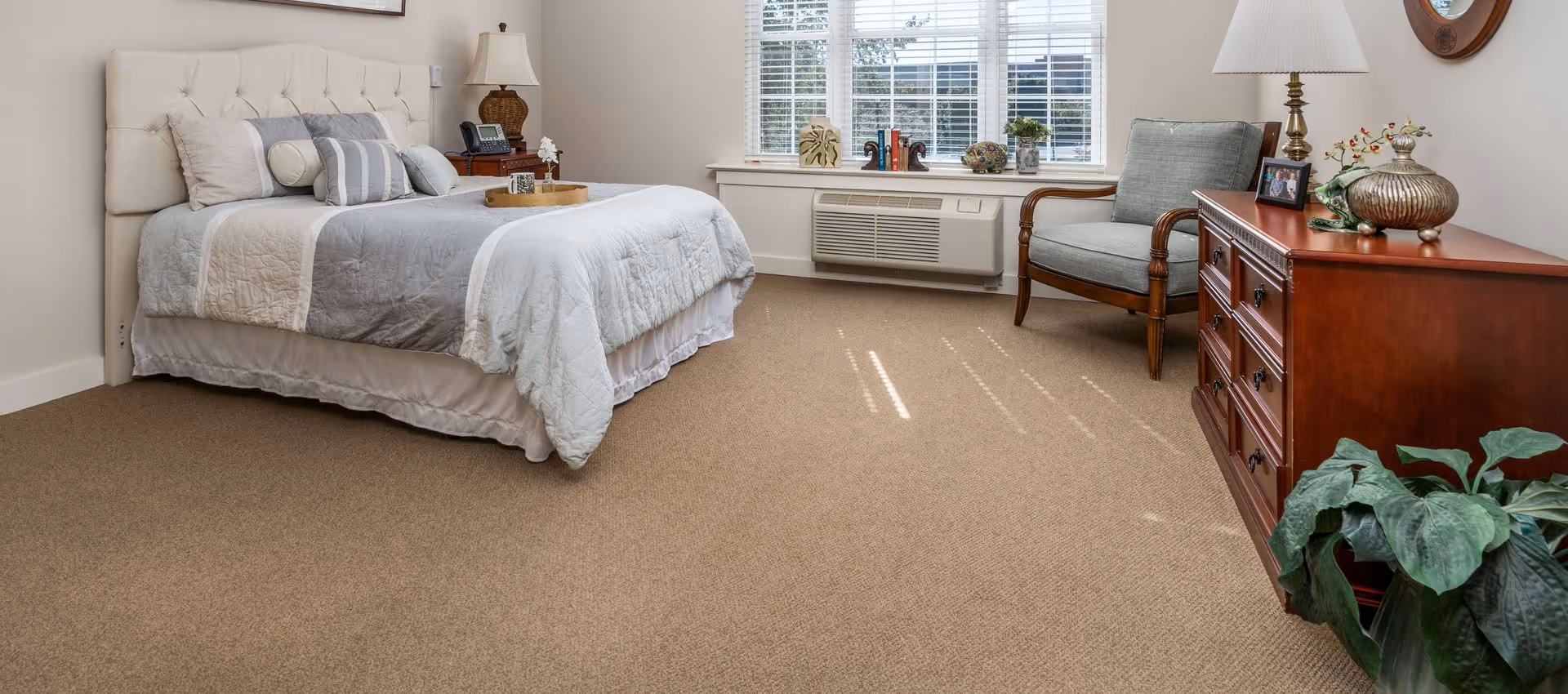Bright, neatly furnished bedroom with a bed, armchair, dresser, window and beige carpet.