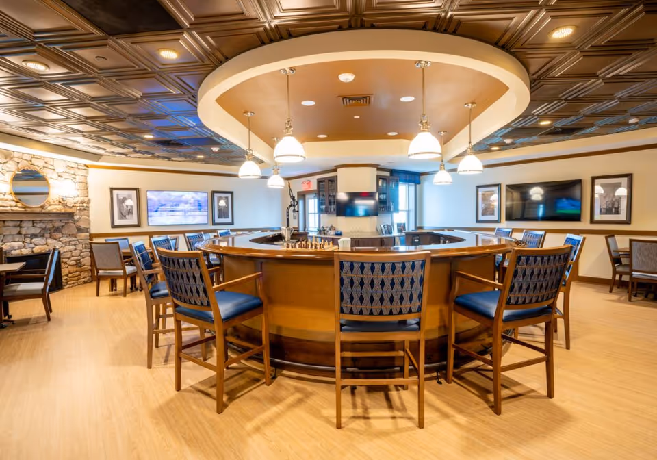 A spacious common area with a large circular wooden table surrounded by chairs with blue patterned cushions. The ceiling features decorative panels and hanging pendant lights. The room has a stone fireplace on the left wall, framed pictures, and two flat-screen TVs mounted on the walls. The floor is light wood, and the overall atmosphere is warm and inviting.