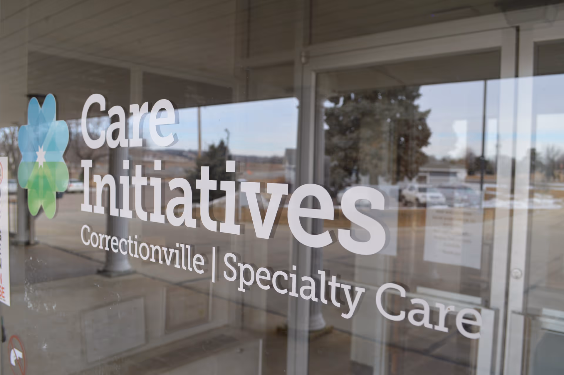 Glass entrance door with the text 'Care Initiatives Correctionville | Specialty Care' printed on it, reflecting the outdoor parking area and trees.