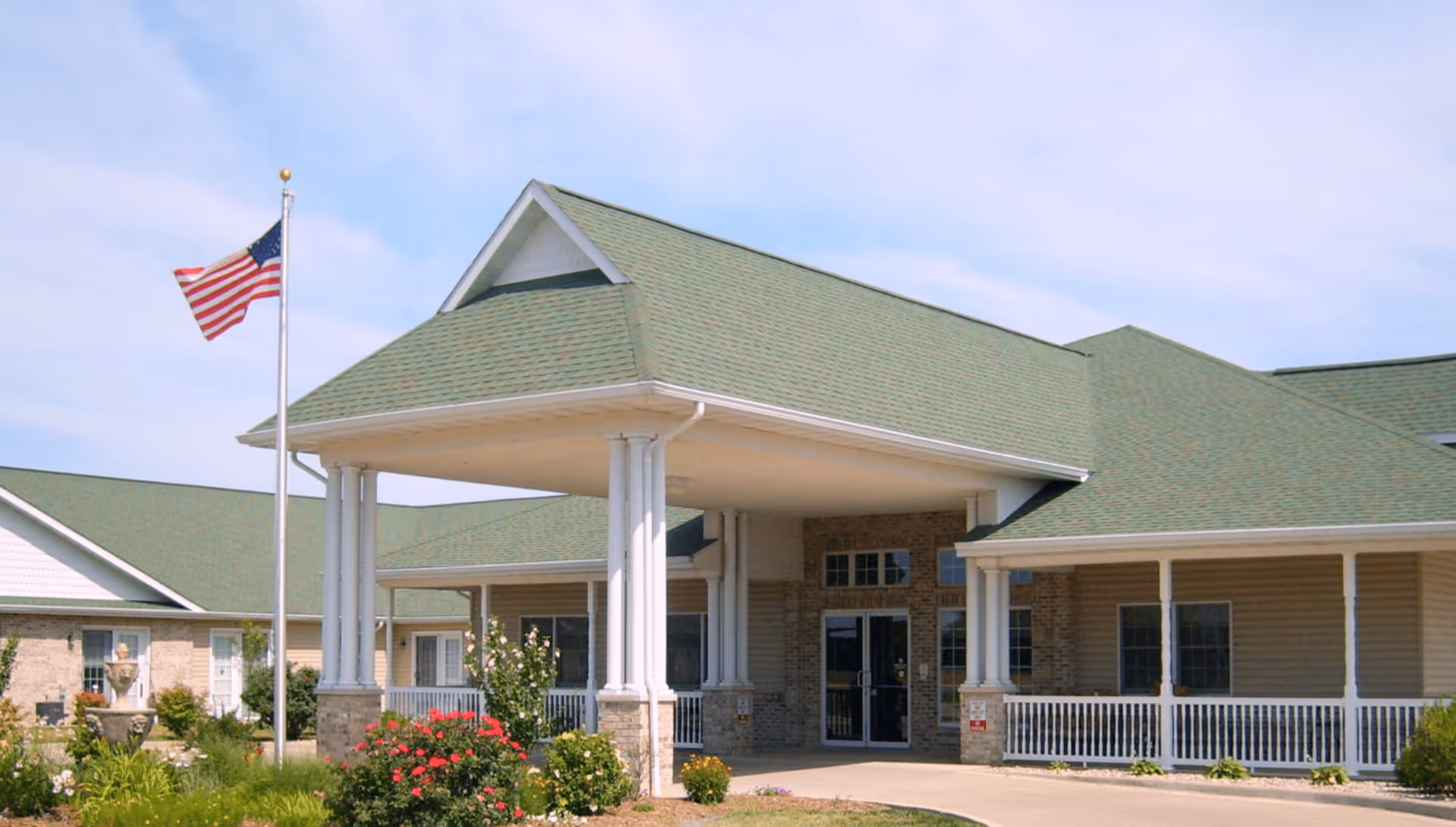 Front exterior view of The Glenwood Supportive Living of Staunton building with a covered entrance supported by white columns, green roof, American flag on a flagpole, and landscaped garden with flowers and shrubs.