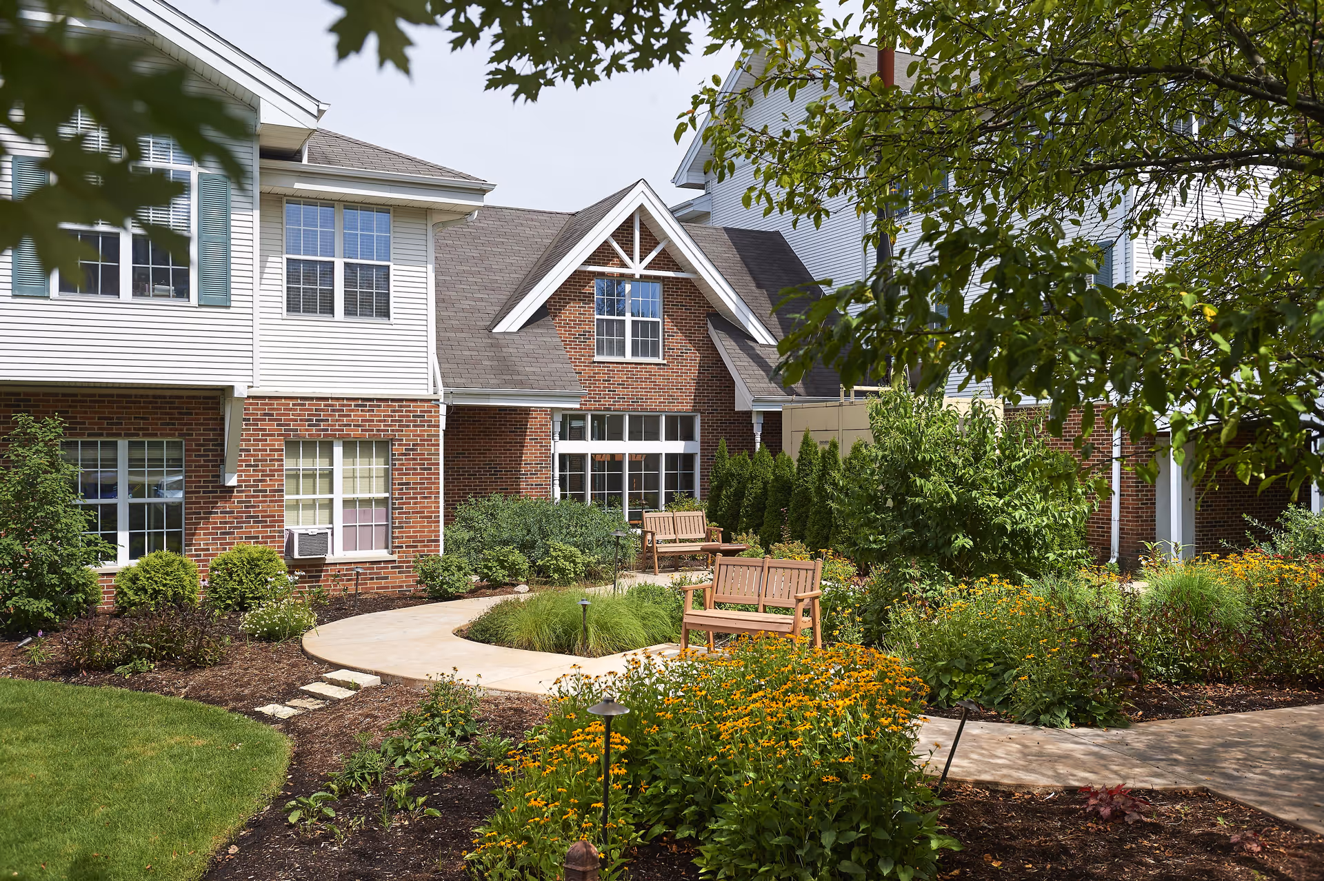 Outdoor garden area at Peace Village featuring a paved walkway, wooden benches, various green shrubs, and yellow flowers, with a two-story building with brick and white siding in the background partially shaded by tree branches.