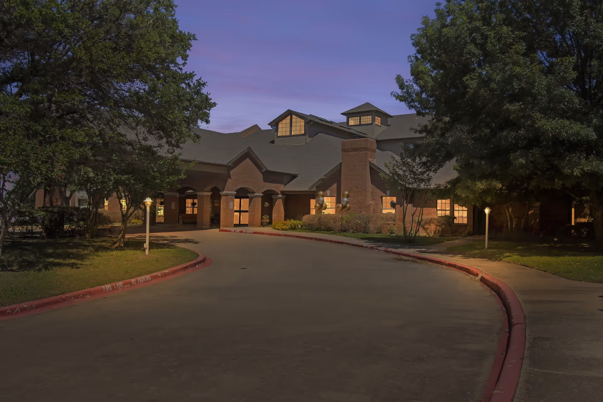 Exterior view of a senior living facility building at dusk with warm lights glowing from the windows. The building has a brick facade, multiple roof peaks, and a covered entrance with arches. Trees and landscaping surround the driveway leading up to the entrance.
