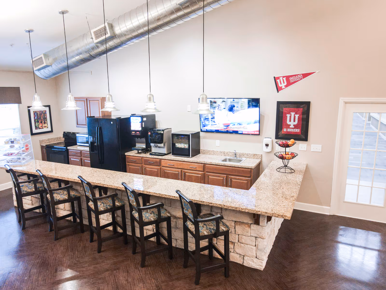 A modern kitchen area with a long granite countertop bar and six high chairs with patterned cushions. The kitchen features wooden cabinets, a black refrigerator, a microwave, a coffee machine, a small beverage cooler, and a sink. Above the counter, five pendant lights hang from the ceiling. On the wall, there is a mounted flat-screen TV showing a program, and two Indiana University decorations. A fruit basket with apples and oranges is placed on the counter near a door with glass panes.