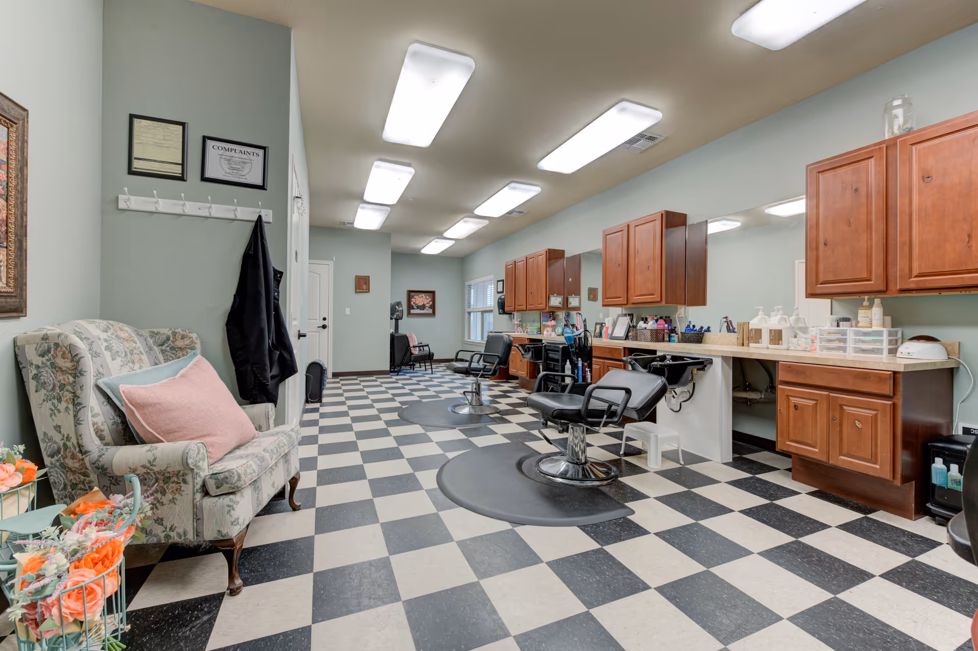 Interior view of a senior living facility's hair salon with checkered black and white flooring, two black salon chairs in front of sinks and wooden cabinets, a floral upholstered armchair with a pink pillow on the left, and bright overhead lighting.