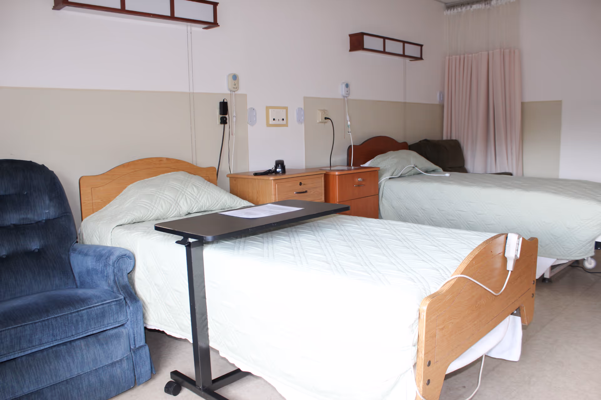 A room with two hospital-style beds covered with light green quilts, each equipped with a call button and an overbed table. There is a blue upholstered chair next to one bed, a wooden nightstand between the beds, and a window with closed curtains in the background.