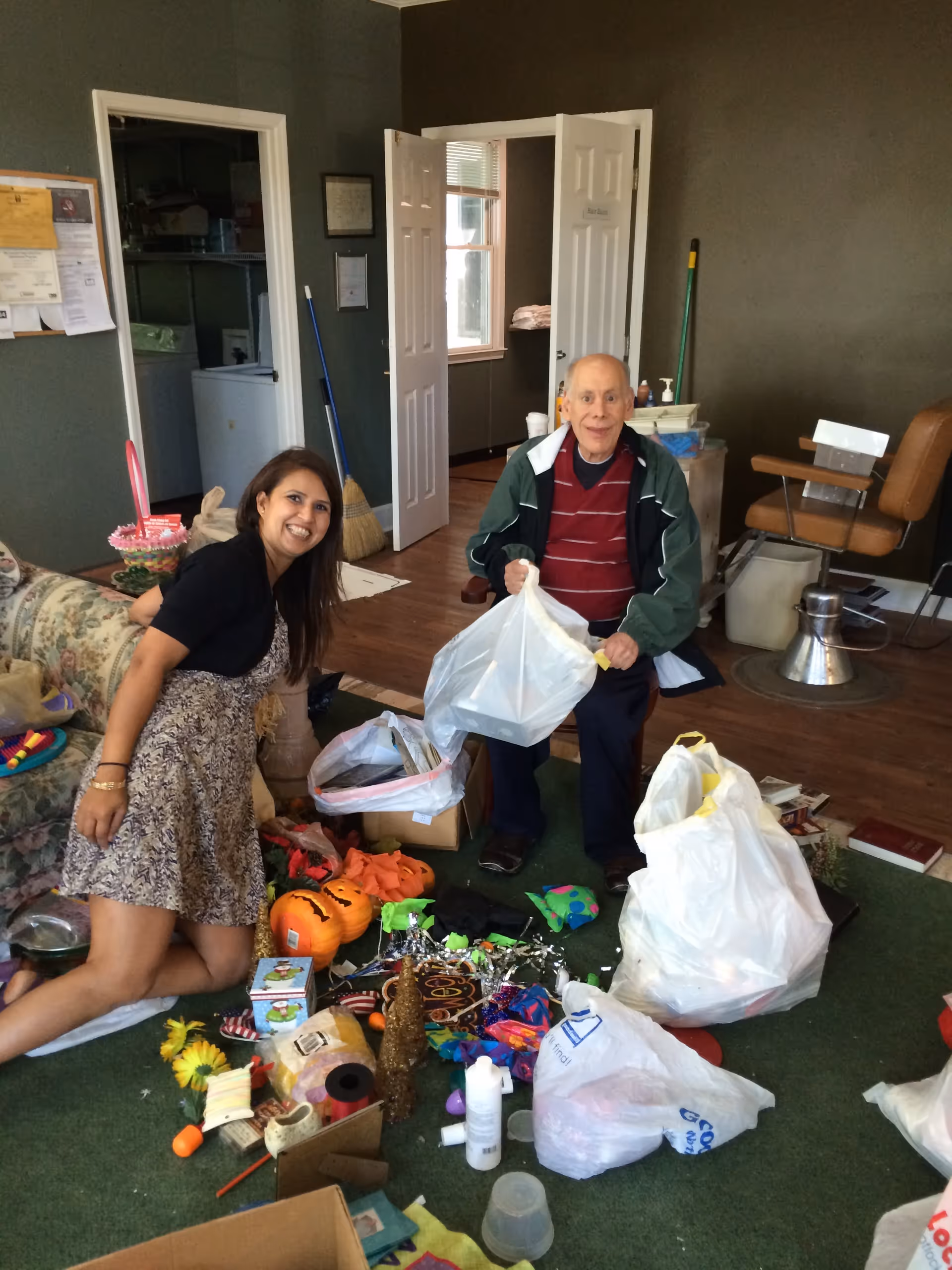 An elderly man sitting on a chair and a woman kneeling on the floor in a room with various items and bags scattered on a green carpet. The room has dark walls, wooden flooring, and an open door leading to another room. There is a floral couch on the left and a brown chair on the right.