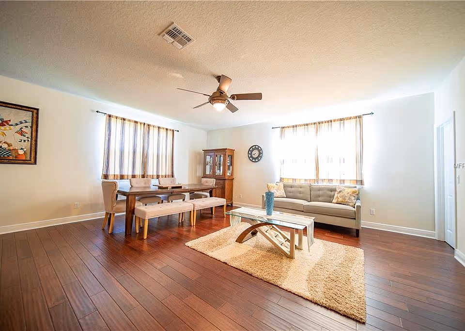 A bright living room with wooden flooring featuring a gray sofa with patterned cushions, a glass coffee table on a beige rug, a wooden dining table with chairs and benches, a wooden cabinet, two windows with striped curtains, a ceiling fan with light, and a framed artwork on the wall.