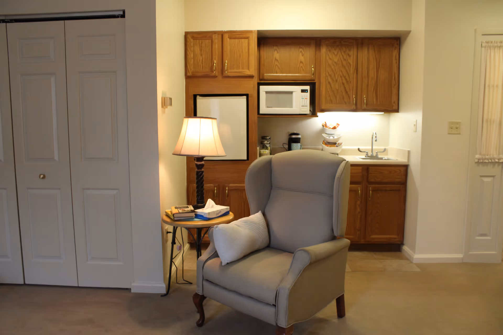 A cozy living space with a wingback armchair and side table in front of a small kitchenette with wooden cabinets and a microwave.