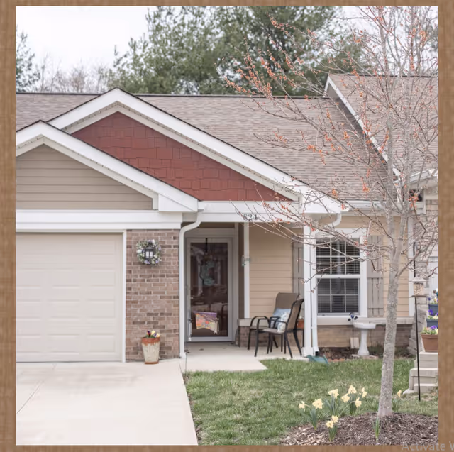 Front exterior of a single-story senior living unit with a garage, small porch with chairs, potted plants and a tree in the yard.