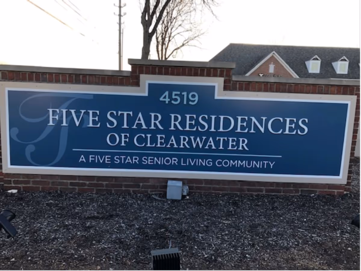 A blue and white sign mounted on a brick base that reads '4519 FIVE STAR RESIDENCES OF CLEARWATER A FIVE STAR SENIOR LIVING COMMUNITY' with a tree and building roof visible in the background.