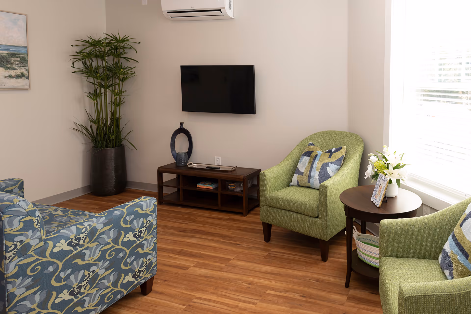 A cozy living room with two green armchairs and one blue patterned armchair arranged around a wooden side table with a vase of white flowers and a framed photo. A flat-screen TV is mounted on the wall above a wooden media console. A tall potted plant is placed in the corner, and a window with blinds allows natural light to brighten the room.