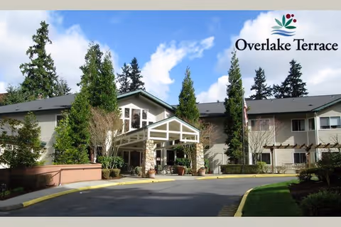 Exterior view of Overlake Terrace Assisted & Senior Living facility showing a two-story building with a covered entrance, surrounded by tall trees and landscaping under a partly cloudy sky.