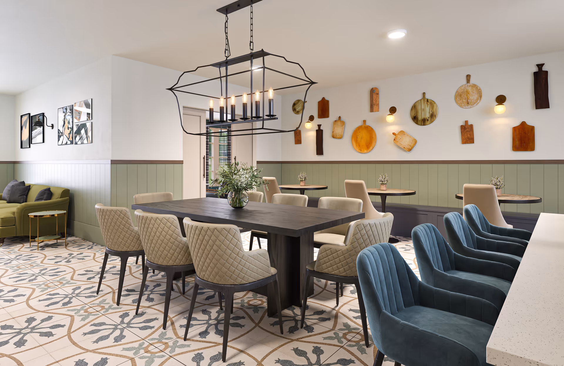 A stylish dining area featuring a large dark wooden table surrounded by beige quilted chairs, a modern black metal chandelier with exposed bulbs hanging above. The room has patterned tile flooring, green wainscoting on the walls, and decorative wooden cutting boards mounted on the far wall. There are additional small round tables with beige chairs and a green sofa with dark cushions in the corner. Blue upholstered chairs are lined up along a white countertop on the right side.