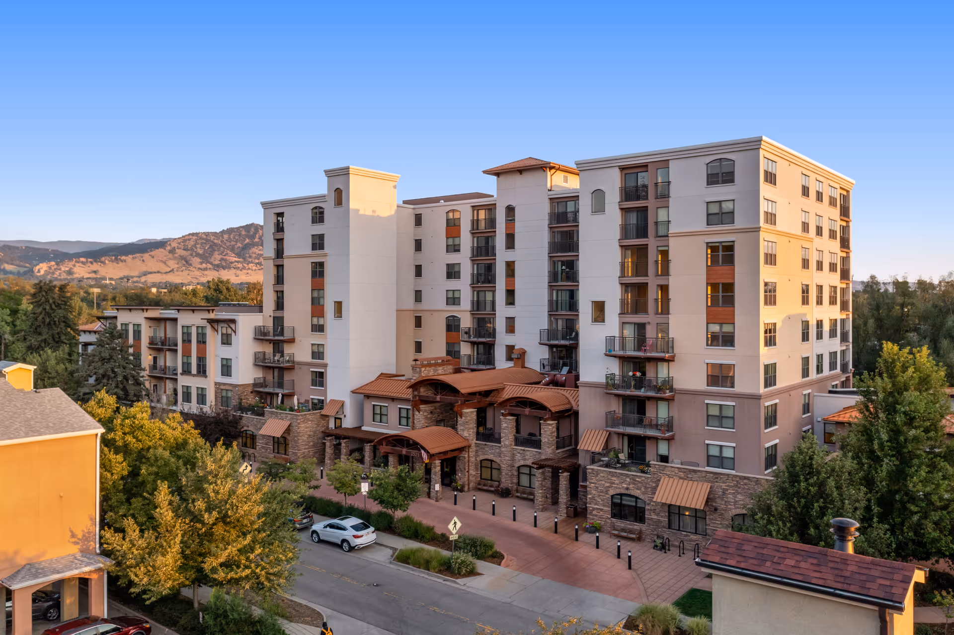 Exterior view of a multi-story senior living facility named The Pearl at Boulder Creek, surrounded by trees and with mountains visible in the background under a clear blue sky.