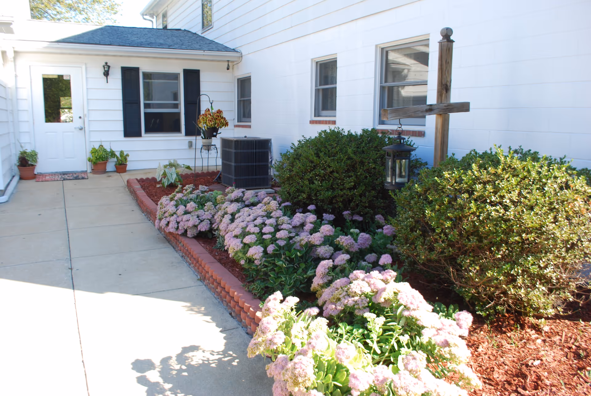 Outdoor garden area at Whitewood Assisted Living with a concrete walkway, flower beds with blooming pink flowers, green shrubs, a hanging lantern on a wooden post, and a white building with windows and a door in the background.