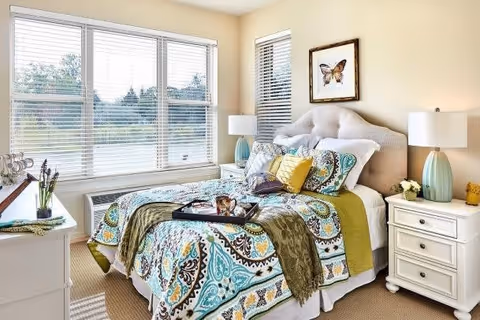 A bright and cozy bedroom with a large window letting in natural light. The bed has a tufted beige headboard and is dressed with a colorful patterned quilt, multiple pillows, and a green throw blanket. There are two white nightstands on either side of the bed, each with a lamp and decorative items. A framed butterfly artwork hangs above the bed.
