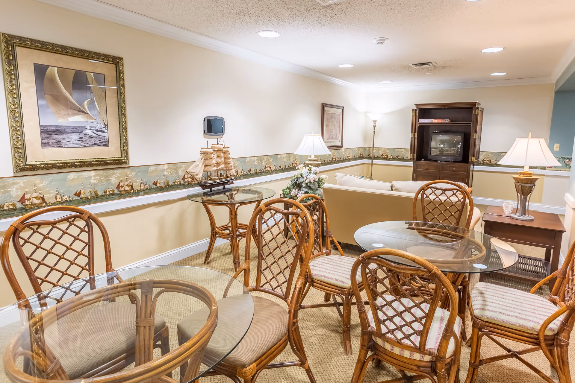 A cozy living room area with a beige couch facing a wooden entertainment center with a TV. In the foreground, there are two glass-top tables surrounded by wicker chairs with cushioned seats. The walls are decorated with a nautical-themed wallpaper border and framed artwork of a sailboat. The room is softly lit by table lamps and a floor lamp.