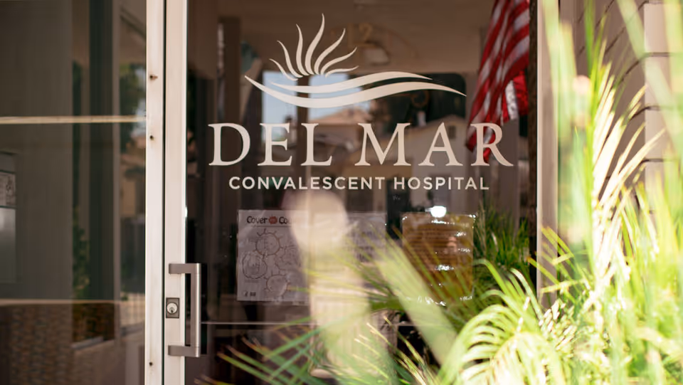 Glass entrance door displaying 'DEL MAR Convalescent Hospital' signage with plants and an American flag visible inside.