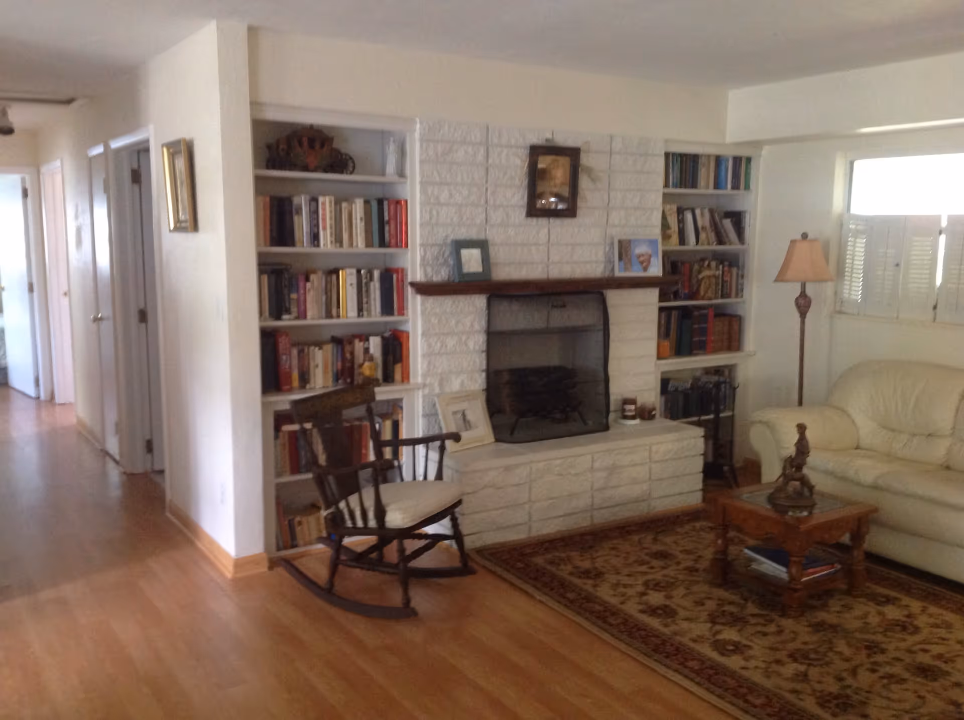 A cozy living room with a white brick fireplace centered between two built-in bookshelves filled with books and decorative items. In front of the fireplace is a wooden rocking chair with a cushion. To the right, there is a beige leather sofa next to a floor lamp and a window with white shutters. A wooden coffee table with a decorative sculpture sits on a patterned area rug on the wooden floor.