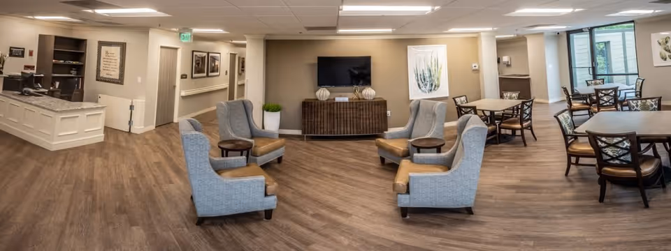 A spacious senior living common area featuring four gray armchairs arranged around two small round tables in the center. A large flat-screen TV is mounted on the wall above a wooden console table with decorative vases. To the right, there are multiple dining tables with chairs, and large windows letting in natural light. On the left side, there is a reception desk with granite countertop and shelves behind it. The room has wood flooring and neutral-colored walls with framed artwork.
