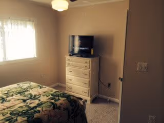 A bedroom with a bed covered in a floral patterned bedspread, a window with sheer curtains allowing natural light, a white chest of drawers with a flat-screen TV on top, and beige walls and carpet.