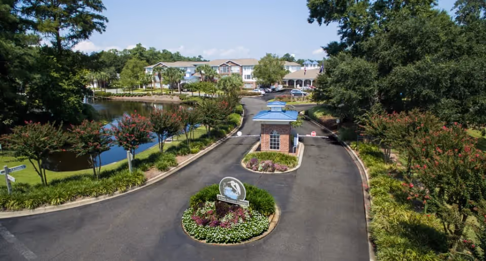 Entrance driveway to The Lakes at Litchfield senior living facility with a small guardhouse in the center, surrounded by landscaped greenery and flowers, a pond on the left, and residential buildings in the background under a blue sky.