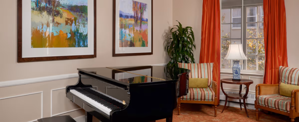 A cozy interior room featuring a black grand piano with a matching bench, two striped armchairs with yellow cushions, a small round wooden table with a decorative lamp, two colorful abstract paintings on the wall, a tall green plant, and a window with orange curtains letting in natural light.