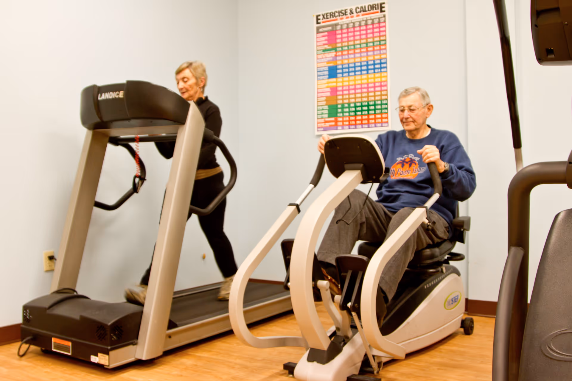 An elderly woman walking on a treadmill and an elderly man using a recumbent exercise bike in a small fitness room with light blue walls and a colorful exercise and calorie chart on the wall.