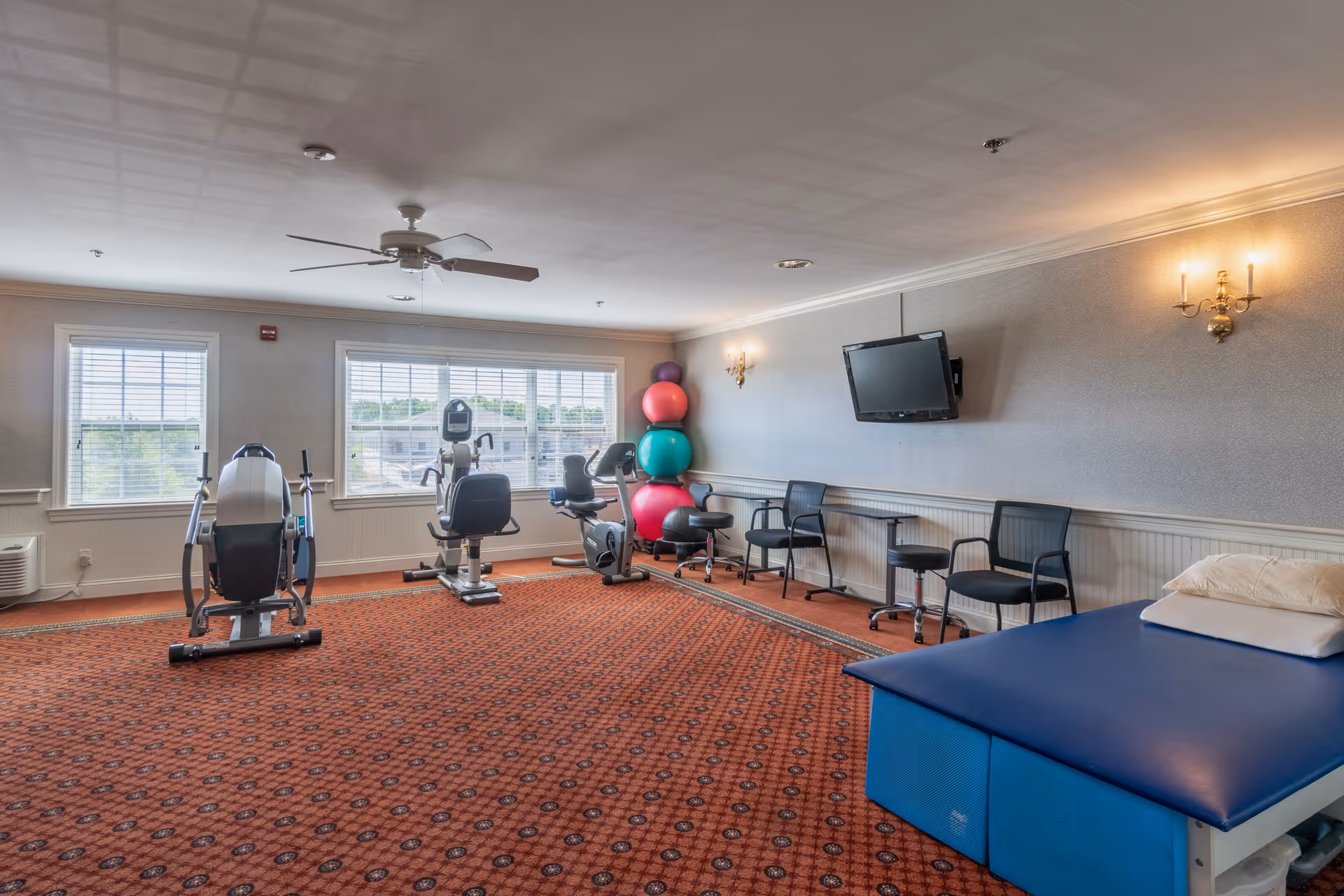 A fitness room in a senior living facility with exercise equipment including stationary bikes and an elliptical machine. There are colorful exercise balls stacked in the corner, a blue padded therapy table with pillows, several chairs along the wall, a wall-mounted TV, and large windows letting in natural light.