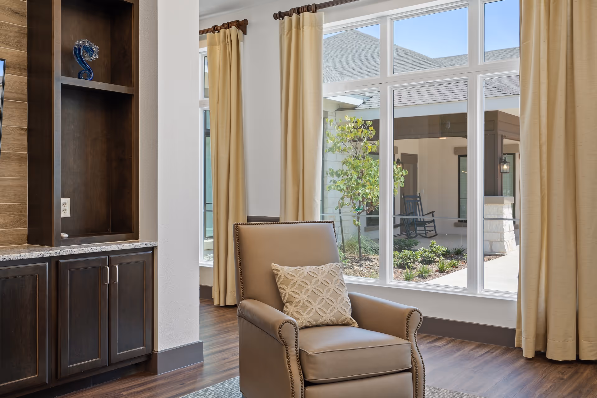A cozy interior corner of a senior living facility featuring a beige armchair with a patterned cushion, positioned near large windows with beige curtains. Outside the windows, there is a view of a courtyard with a small tree, shrubs, and a covered porch with a rocking chair. Inside, there is a dark wood built-in cabinet with granite countertop and a decorative glass sculpture on the shelf.