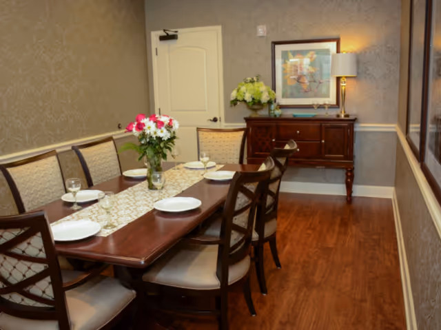 A formal dining room with a wooden dining table set for six people. The table has a decorative runner, a vase with pink and white flowers, and six place settings with plates and wine glasses. The room features patterned wallpaper, a wooden sideboard with a lamp, a framed artwork above it, and a floral arrangement. The floor is wooden, and there is a white door in the background.