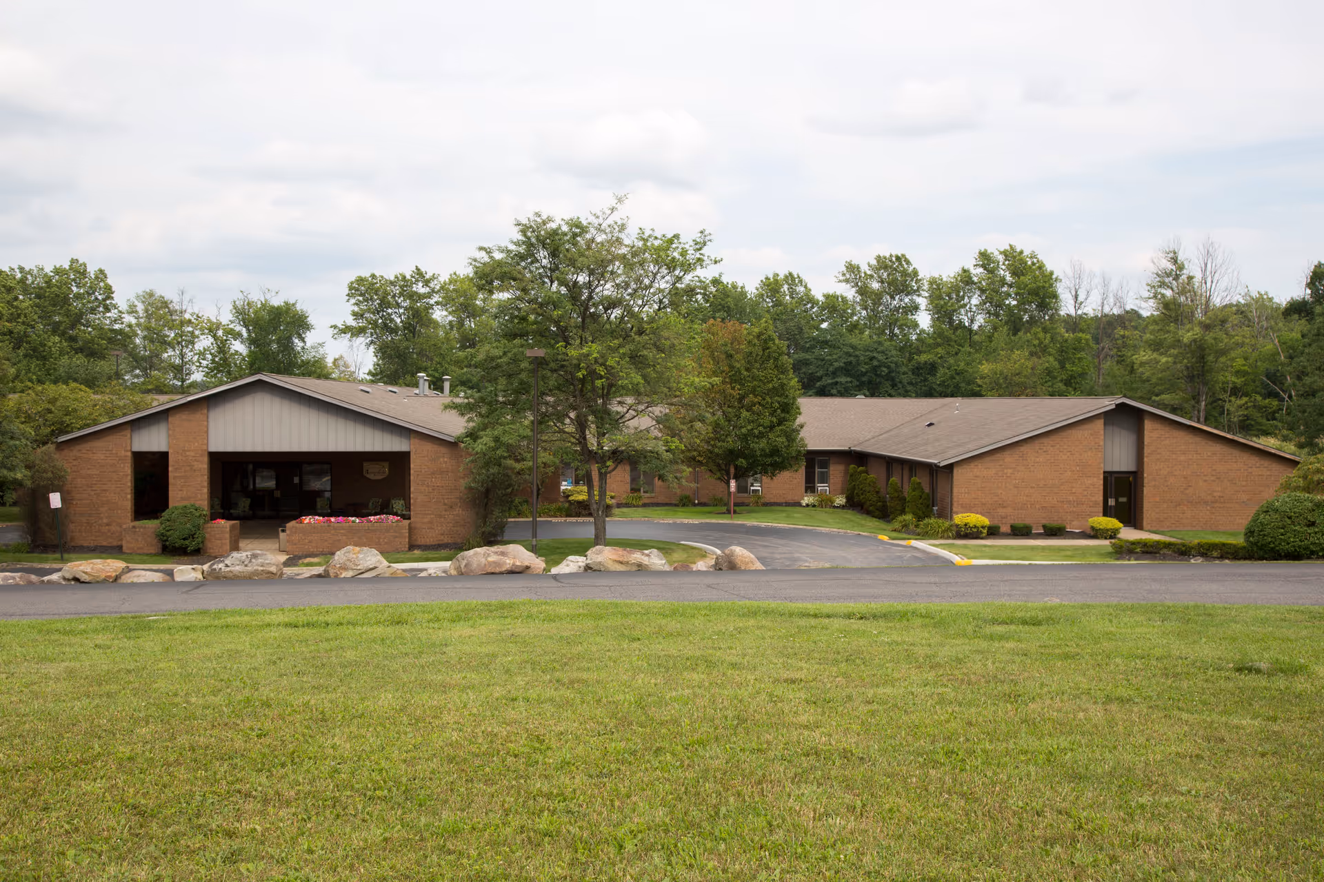 Front exterior view of a single-story brick building with a driveway and landscaped greenery including trees, bushes, and a grassy lawn under a cloudy sky.