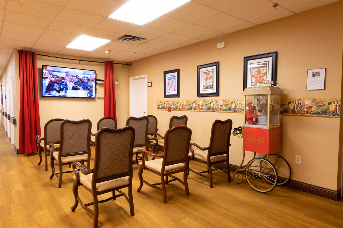 Upholstered chairs arranged facing a wall-mounted TV in a cozy activity/common room with framed posters and a popcorn cart.