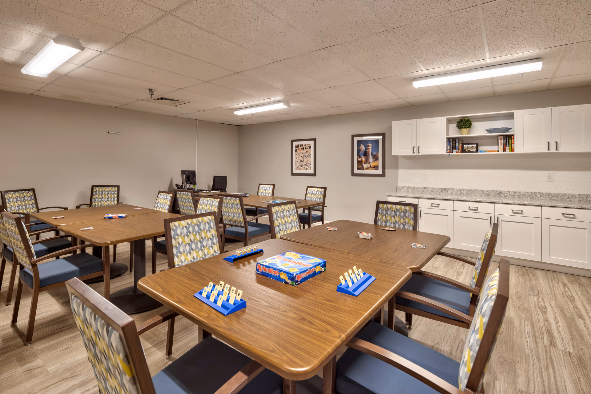 A well-lit activity room with several wooden tables and patterned chairs arranged around them. On the nearest table, there is a Rummikub game set up with tiles and racks. The room has light-colored walls, a wood-patterned floor, white cabinets with shelves holding books and decorative items, and framed pictures on the wall.