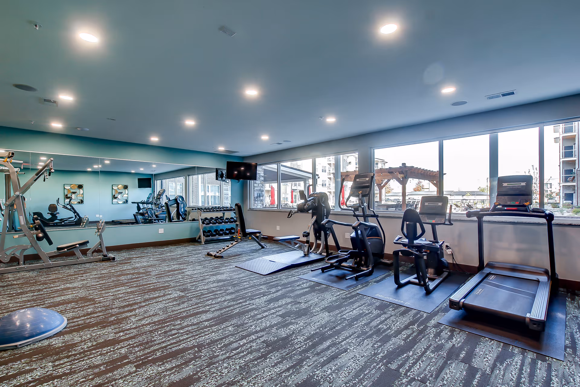 Well-lit fitness room with treadmills, exercise bikes, weight equipment and a mirrored wall next to large windows.