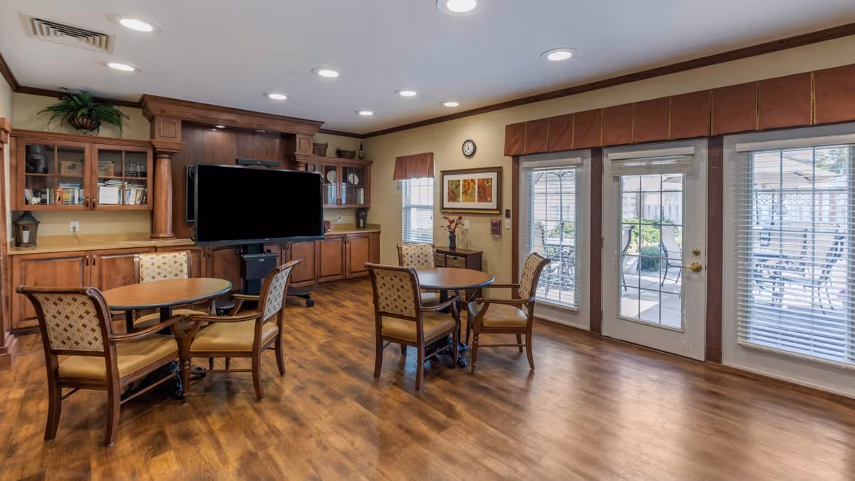 A cozy common area with wooden flooring, two round tables each surrounded by four cushioned chairs, a large flat-screen TV mounted on a wooden cabinet with shelves, and large windows and glass doors leading to an outdoor patio with seating.