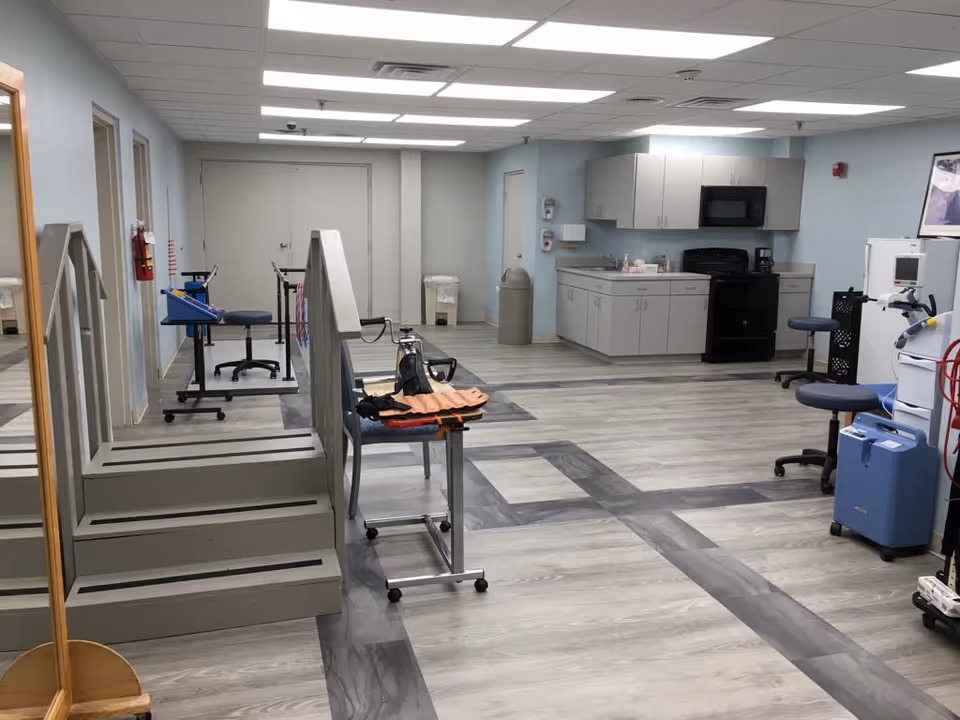 A rehabilitation room with a small set of stairs with handrails for physical therapy, exercise equipment, chairs, and a kitchenette area with cabinets, a stove, and a microwave. The room has light blue walls and wood-patterned flooring.