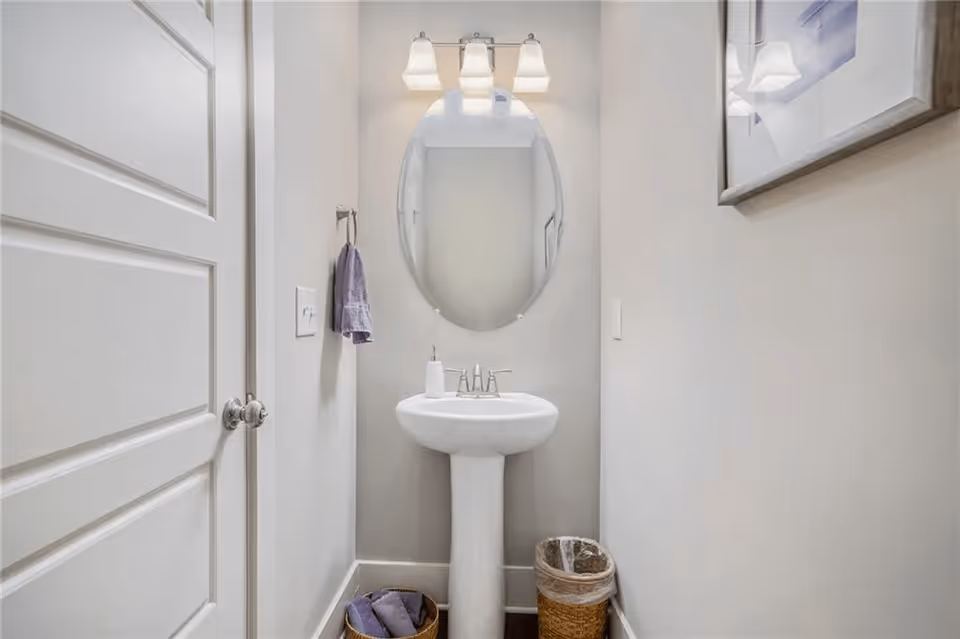 Small bathroom with a white pedestal sink, an oval mirror above it, a three-light fixture, a hand towel hanging on the wall, a wastebasket, and a basket with rolled towels on the floor.
