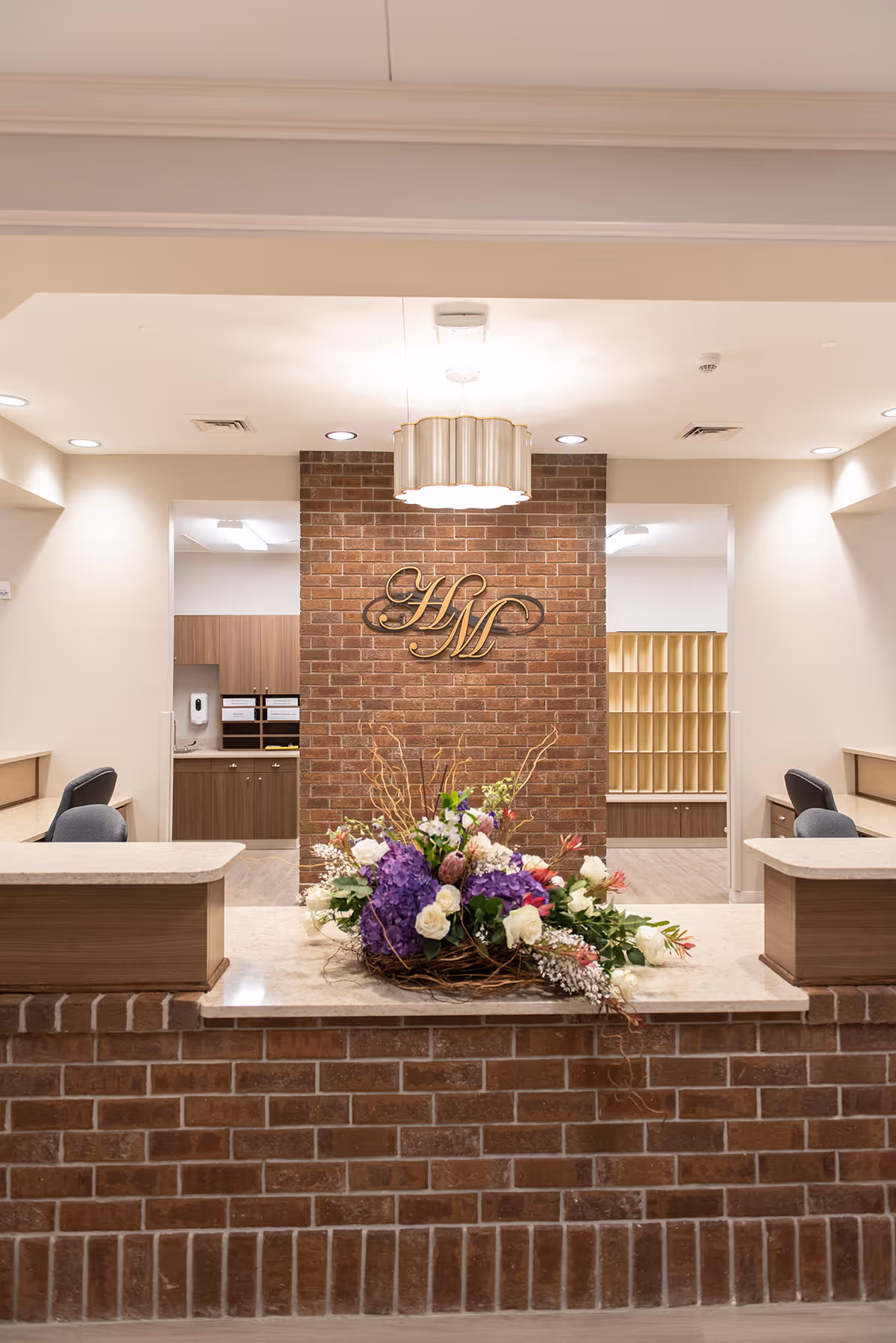 Reception desk and lobby area featuring a brick central wall with a gold "HM" logo, a hanging light, and a flower arrangement on the counter.