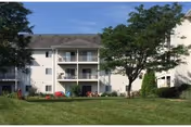 Exterior view of a two-story senior apartment building with balconies, surrounded by green grass, trees, and shrubs under a clear blue sky.