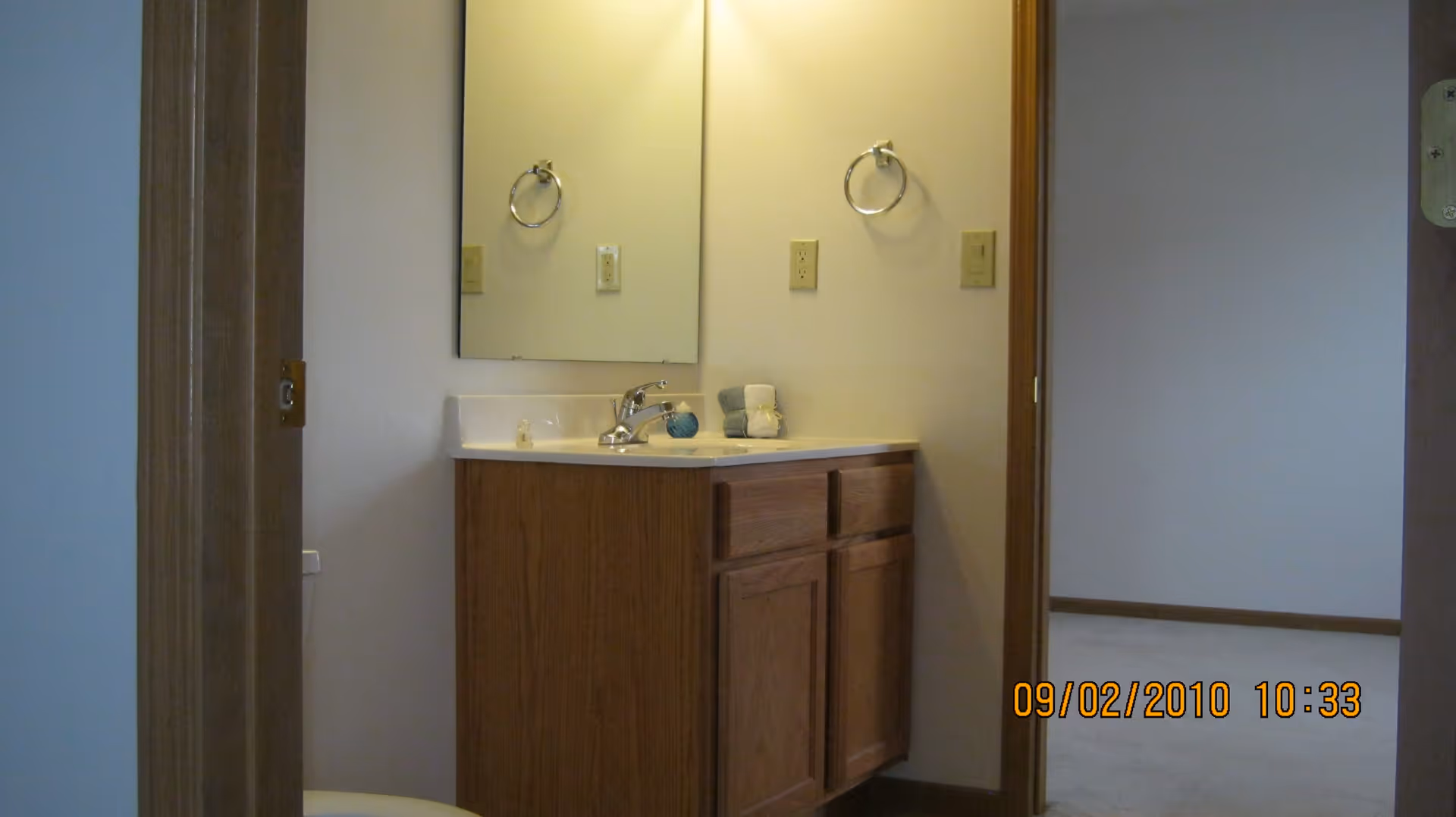 A small bathroom vanity with a wooden cabinet and a white countertop with a faucet. Above the vanity is a rectangular mirror mounted on the wall. A towel ring is mounted on the wall to the right of the mirror. The bathroom door is open, showing part of an adjacent room with carpeted flooring.