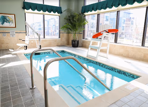 Sunlit indoor therapy pool with stainless handrails, a pool lift chair, large windows and a potted plant.