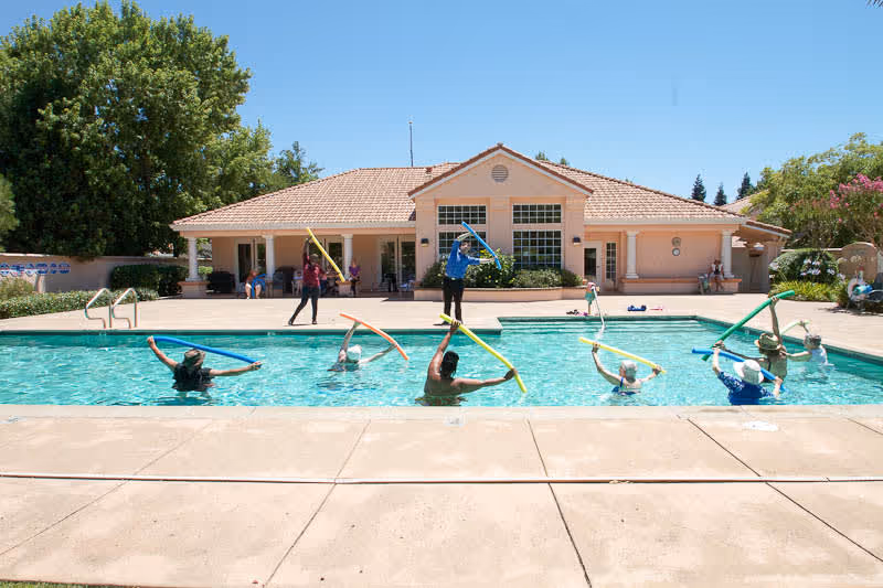 A group of seniors participating in a water exercise class in an outdoor swimming pool at a senior living facility. The pool is surrounded by a concrete deck, and a building with a tiled roof and large windows is visible in the background. Trees and shrubs surround the area under a clear blue sky.