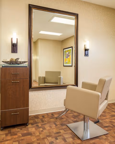 A beige salon chair in front of a large wall-mounted mirror with a dark wooden frame. On the left side, there is a wooden cabinet with three drawers and a decorative bowl on top. The walls are light-colored with two modern wall sconces on either side of the mirror. The floor has a parquet wood pattern.