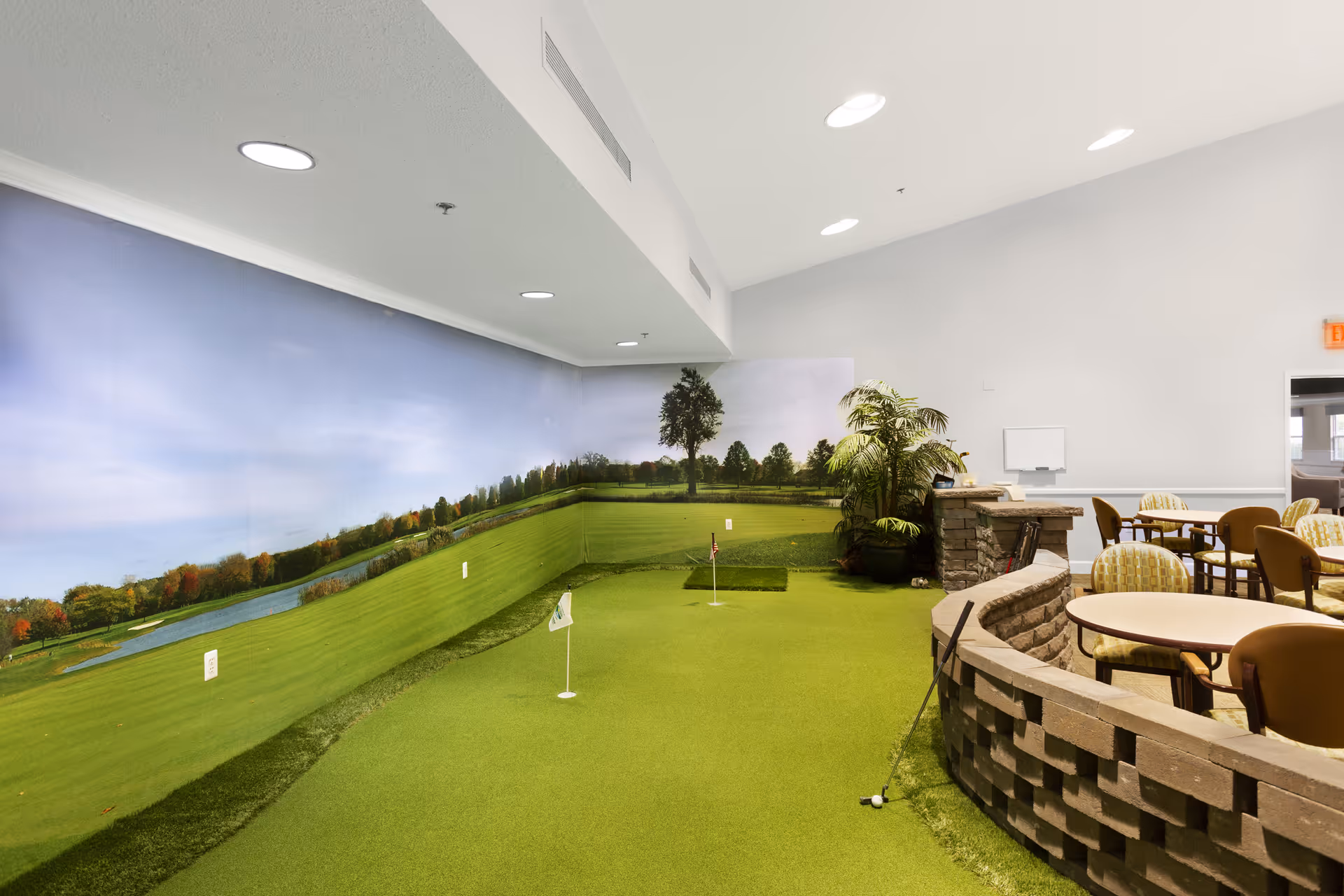 Indoor putting green with a golf-course mural and seating tables and chairs in a senior living common area.