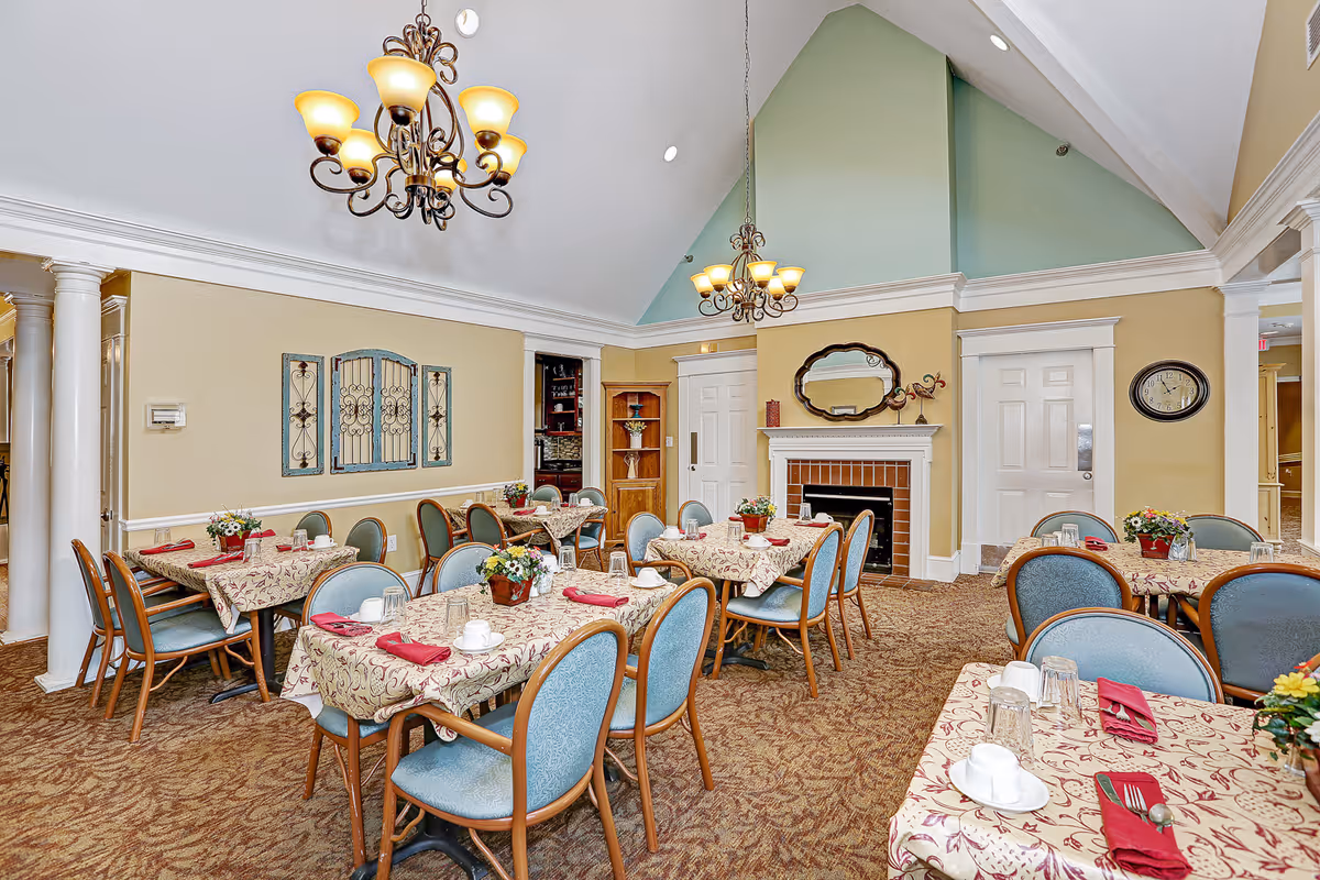 A dining room with multiple tables covered in patterned tablecloths, each set with red napkins, white cups, and glasses. The room features blue cushioned chairs, a fireplace with a decorative mirror above it, two ornate chandeliers hanging from a vaulted ceiling, and light yellow walls with white trim.