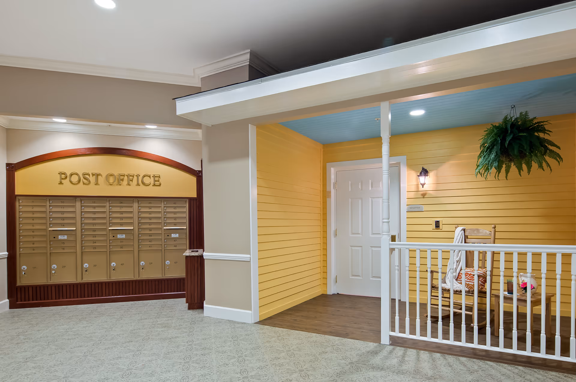 Indoor area with a section designed like a small porch featuring yellow walls, a white door, a wooden rocking chair with a blanket and pillow, a small table with a flower vase, and a hanging plant. To the left, there is a wall of mailboxes labeled 'POST OFFICE' in gold letters.