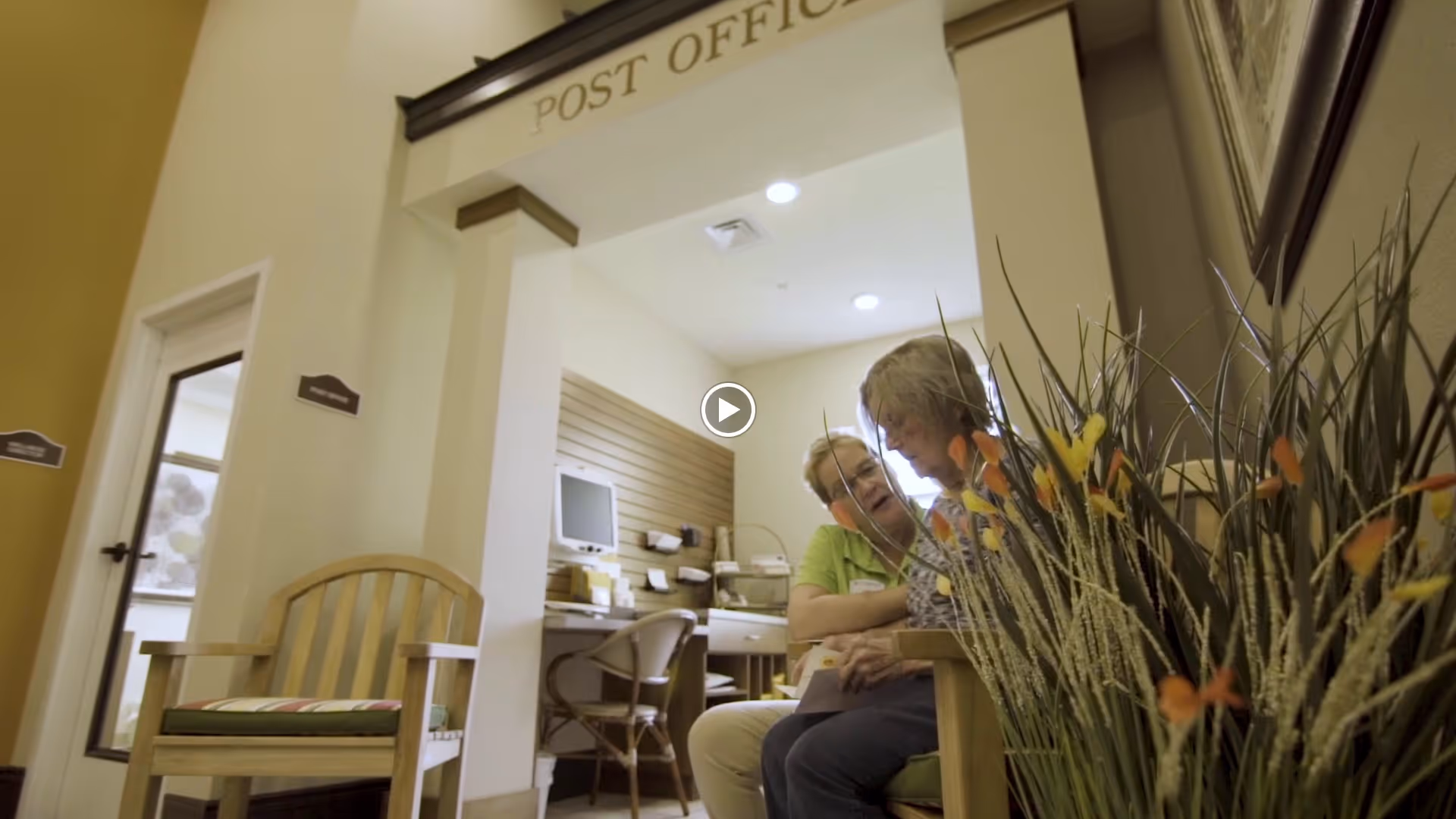 Two elderly women sitting and talking in a small room labeled 'Post Office' inside a senior living facility. The room has a desk with a computer and mail slots on the wall. There is a wooden chair with a cushion in the foreground and decorative plants on the right side.